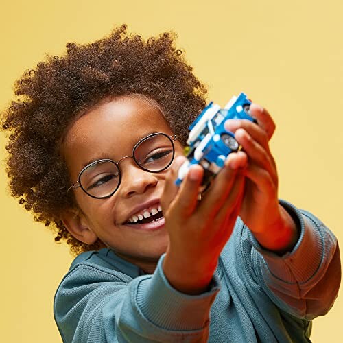 Child with glasses smiling and holding a toy car against a yellow background.